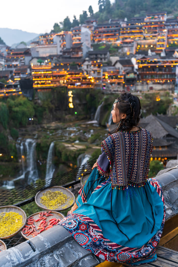 Vista desde atrás de una mujer sentada en un tejado con un vestido azul y chal bordado tradicional, mirando hacia un pintoresco pueblo de montaña chino iluminado con luces doradas, con cascadas que caen entre la vegetación y cestas de secado en primer plano.