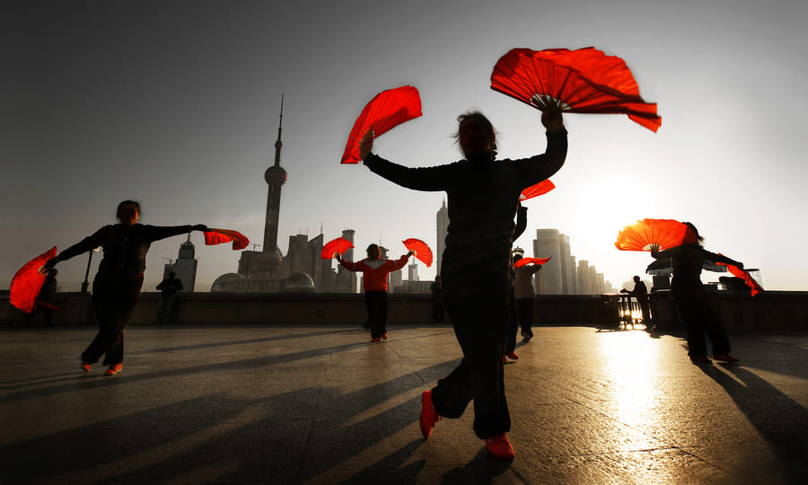 Siluetas de personas practicando una danza tradicional china con grandes abanicos rojos en una plaza pública al amanecer, con el horizonte urbano de rascacielos de Shanghái de fondo bajo una luz solar brillante y sombras alargadas en el suelo.