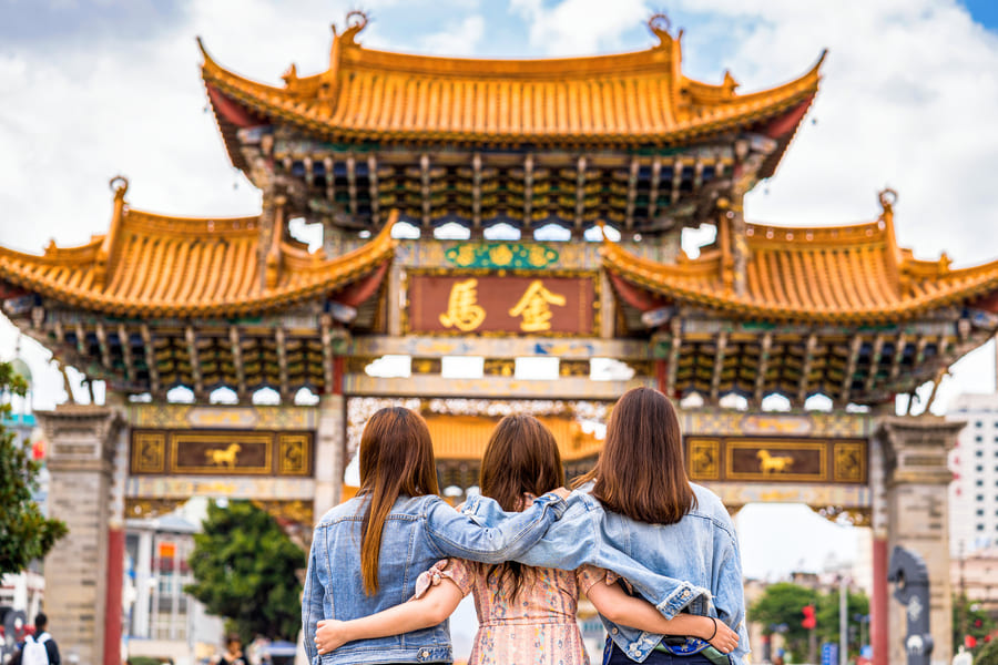 Tres jóvenes mujeres con chaquetas de mezclilla y los brazos sobre los hombros, vistas desde atrás, admirando la ornamentada puerta histórica 'Golden Horse' (Jinma Biji) en Kunming, China, con su arquitectura tradicional y un cartel con caracteres chinos.