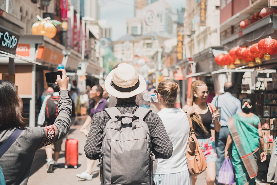 Grupo de turistas con mochilas recorriendo a pie una concurrida calle comercial asiática decorada con faroles chinos rojos; una muestra de la inmersión cultural en nuestros viajes personalizados a China.